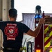 A firefighter wearing a navy blue 'Lajes Field Bombeiros' t-shirt is seen from behind, directing or guiding a fire truck out of a fire station bay. The shirt displays the 765th ABS/CEF unit insignia with a bulldog mascot and the nickname 'Fire Dawgs', indicating a U.S. Air Force fire department unit stationed at Lajes Field in the Azores, Portugal. The firefighter is gesturing with one hand pointing outward while the other is raised against a wall.
