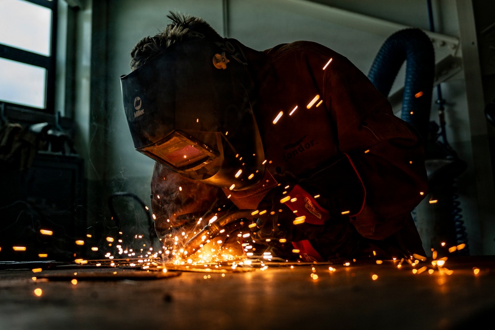 A welder is working close to the ground in an industrial workshop, producing a dramatic shower of bright orange sparks. The worker is wearing a Miller-brand welding helmet and protective orange/brown leather gear. The low-angle shot captures the intensity of the metalworking process with sparks scattered across the floor.