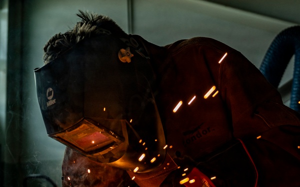 A welder is working close to the ground in an industrial workshop, producing a dramatic shower of bright orange sparks. The worker is wearing a Miller-brand welding helmet and protective orange/brown leather gear. The low-angle shot captures the intensity of the metalworking process with sparks scattered across the floor.