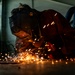 A welder is working close to the ground in an industrial workshop, producing a dramatic shower of bright orange sparks. The worker is wearing a Miller-brand welding helmet and protective orange/brown leather gear. The low-angle shot captures the intensity of the metalworking process with sparks scattered across the floor.