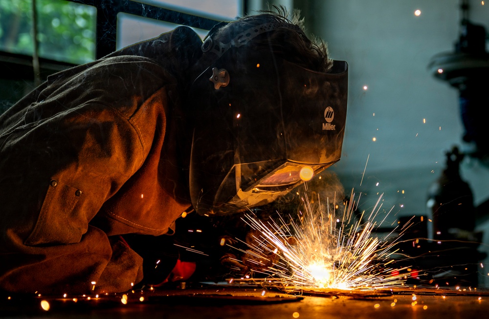 A welder is captured in close-up while performing a welding operation on a metal surface at floor level. The worker is wearing a Miller-brand auto-darkening welding helmet and a brown leather welding jacket, with bright golden sparks radiating outward from the weld point. The image is dramatically lit by the welding arc, creating a high-contrast, visually striking industrial scene.