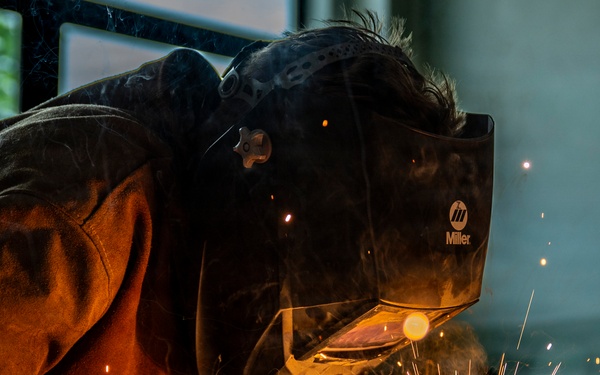 A welder is captured in close-up while performing a welding operation on a metal surface at floor level. The worker is wearing a Miller-brand auto-darkening welding helmet and a brown leather welding jacket, with bright golden sparks radiating outward from the weld point. The image is dramatically lit by the welding arc, creating a high-contrast, visually striking industrial scene.