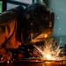 A welder is captured in close-up while performing a welding operation on a metal surface at floor level. The worker is wearing a Miller-brand auto-darkening welding helmet and a brown leather welding jacket, with bright golden sparks radiating outward from the weld point. The image is dramatically lit by the welding arc, creating a high-contrast, visually striking industrial scene.