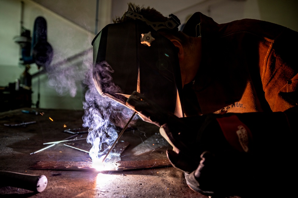A welder is working close to the ground in what appears to be an industrial workshop or garage, performing a welding operation on a metal surface. The scene is dramatically lit by the intense arc light of the welding process, with visible smoke and sparks rising from the work area. The worker is equipped with appropriate protective gear including a welding helmet and leather jacket.