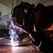 A welder is working close to the ground in what appears to be an industrial workshop or garage, performing a welding operation on a metal surface. The scene is dramatically lit by the intense arc light of the welding process, with visible smoke and sparks rising from the work area. The worker is equipped with appropriate protective gear including a welding helmet and leather jacket.