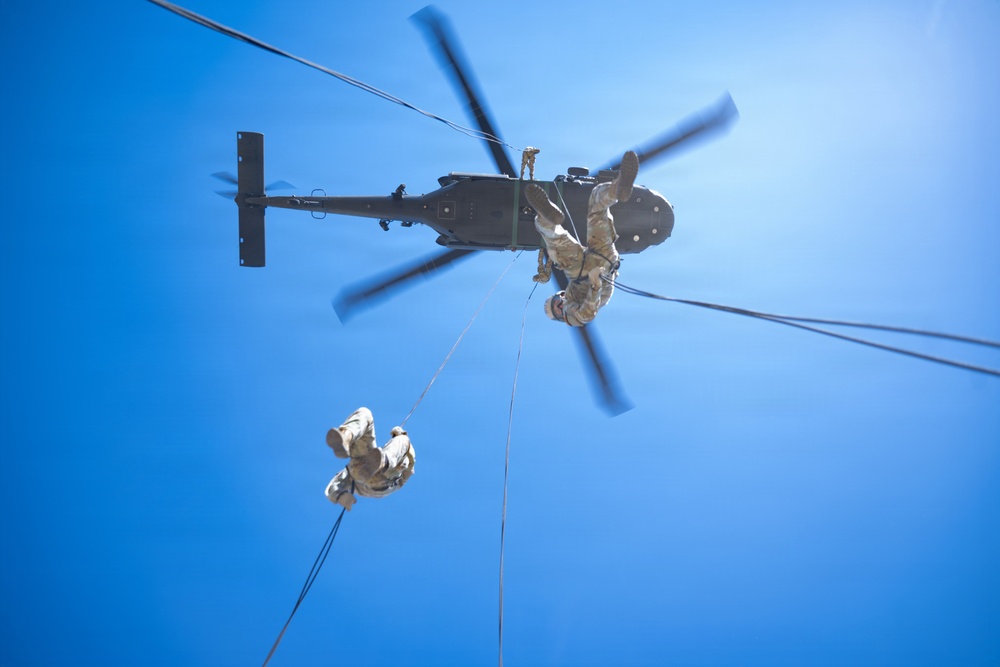 A low-angle upward shot captures military personnel fast-roping or rappelling from a dark-colored military helicopter against a clear blue sky. At least two soldiers in desert/tan camouflage uniforms are descending on ropes from the hovering aircraft. The rotor blades show motion blur, indicating the helicopter is in active flight.