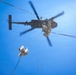 A low-angle upward shot captures military personnel fast-roping or rappelling from a dark-colored military helicopter against a clear blue sky. At least two soldiers in desert/tan camouflage uniforms are descending on ropes from the hovering aircraft. The rotor blades show motion blur, indicating the helicopter is in active flight.