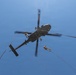 A military helicopter, viewed from directly below, is hovering while personnel rappel down ropes from both sides of the aircraft. The image is taken from a ground-level upward perspective against a clear blue sky. Multiple ropes extend downward from the helicopter with at least two visible personnel in the process of fast-roping or rappelling.