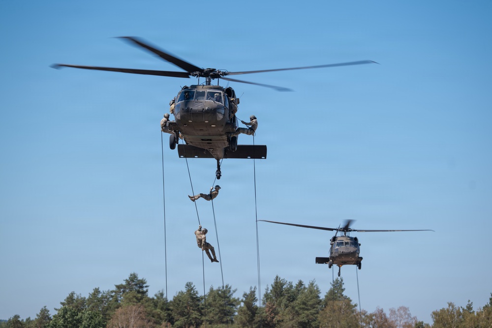 Two military helicopters, identifiable as UH-60 Black Hawks, are shown in flight against a clear blue sky. Personnel are rappelling from ropes suspended beneath the foreground helicopter, while crew members are visible in the open doorways. A second Black Hawk helicopter is visible in the background performing a similar operation.
