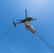 A low-angle upward shot captures a military personnel suspended beneath a helicopter via ropes or rappelling lines against a clear blue sky. The individual appears to be engaged in a fast-rope or rappelling exercise from what appears to be a military utility helicopter. Multiple lines extend outward from the helicopter, suggesting a training or operational insertion/extraction scenario.