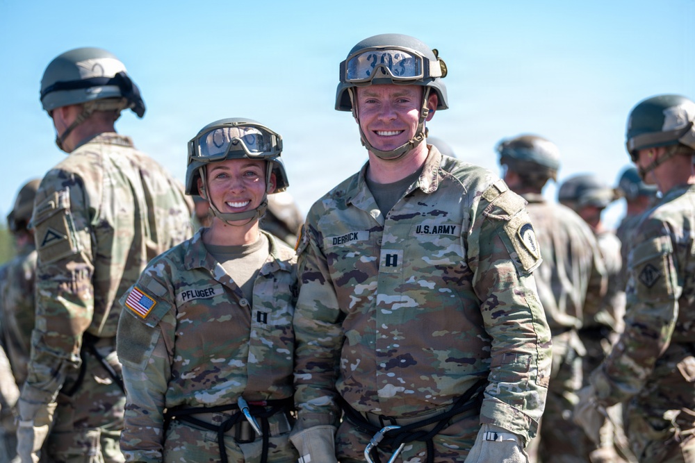 Two U.S. Army soldiers wearing OCP (Operational Camouflage Pattern) uniforms, combat helmets, and ballistic goggles pose together smiling for a photo. Name tapes visible on their uniforms read 'PFLUGER' and 'DERRICK', both appearing to hold the rank of Captain (indicated by the Captain's bars insignia). Additional soldiers in similar gear are visible in the background.