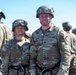 Two U.S. Army soldiers wearing OCP (Operational Camouflage Pattern) uniforms, combat helmets, and ballistic goggles pose together smiling for a photo. Name tapes visible on their uniforms read 'PFLUGER' and 'DERRICK', both appearing to hold the rank of Captain (indicated by the Captain's bars insignia). Additional soldiers in similar gear are visible in the background.