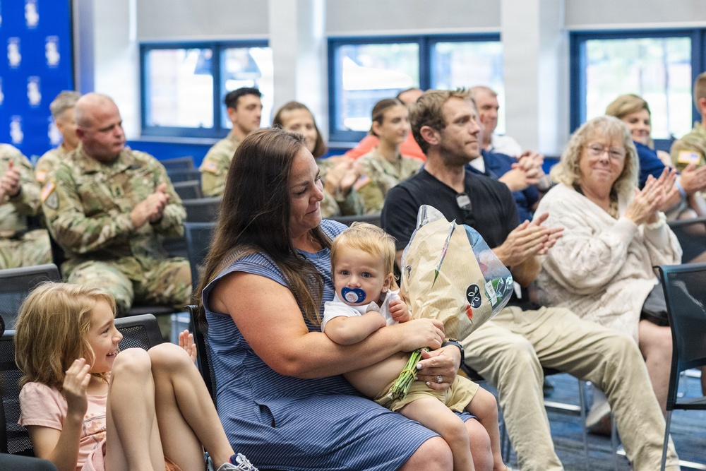 A group of people are seated in a room during what appears to be a military ceremony or recognition event. A woman in a blue dress holds a toddler with a pacifier and a bouquet of flowers, while a young girl beside her smiles and claps. Audience members, including uniformed military personnel in camouflage, are applauding in the background.