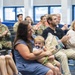 A group of people are seated in a room during what appears to be a military ceremony or recognition event. A woman in a blue dress holds a toddler with a pacifier and a bouquet of flowers, while a young girl beside her smiles and claps. Audience members, including uniformed military personnel in camouflage, are applauding in the background.