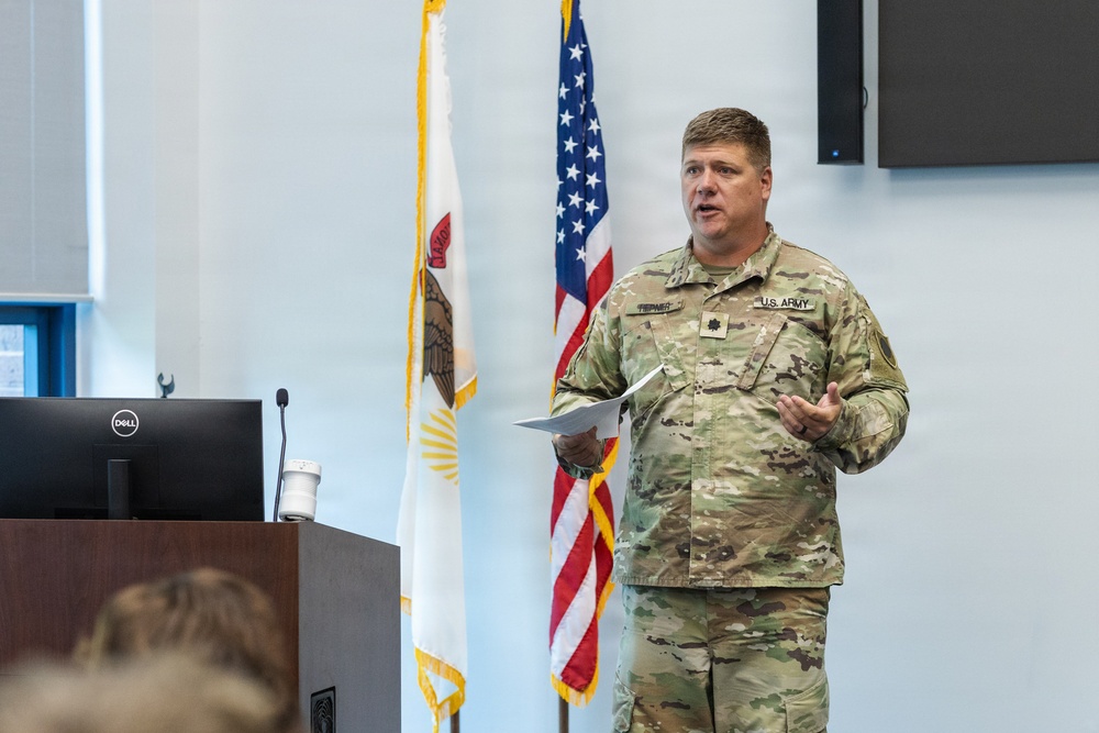 A U.S. Army officer in Operational Camouflage Pattern (OCP) uniform is delivering a presentation or briefing at a podium. He holds papers in one hand and gestures with the other, standing before an American flag and what appears to be an Illinois state flag. The setting appears to be a formal military briefing room or conference facility.