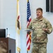 A U.S. Army officer in Operational Camouflage Pattern (OCP) uniform is delivering a presentation or briefing at a podium. He holds papers in one hand and gestures with the other, standing before an American flag and what appears to be an Illinois state flag. The setting appears to be a formal military briefing room or conference facility.