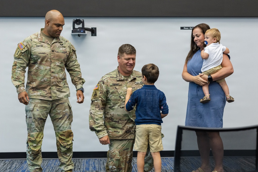 A U.S. Army military ceremony appears to be taking place indoors, likely a promotion or recognition event. Two soldiers in Army Combat Uniforms (ACUs) are interacting with a young boy, while a woman holds an infant nearby. The setting appears to be a conference or briefing room.