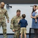 A U.S. Army military ceremony appears to be taking place indoors, likely a promotion or recognition event. Two soldiers in Army Combat Uniforms (ACUs) are interacting with a young boy, while a woman holds an infant nearby. The setting appears to be a conference or briefing room.