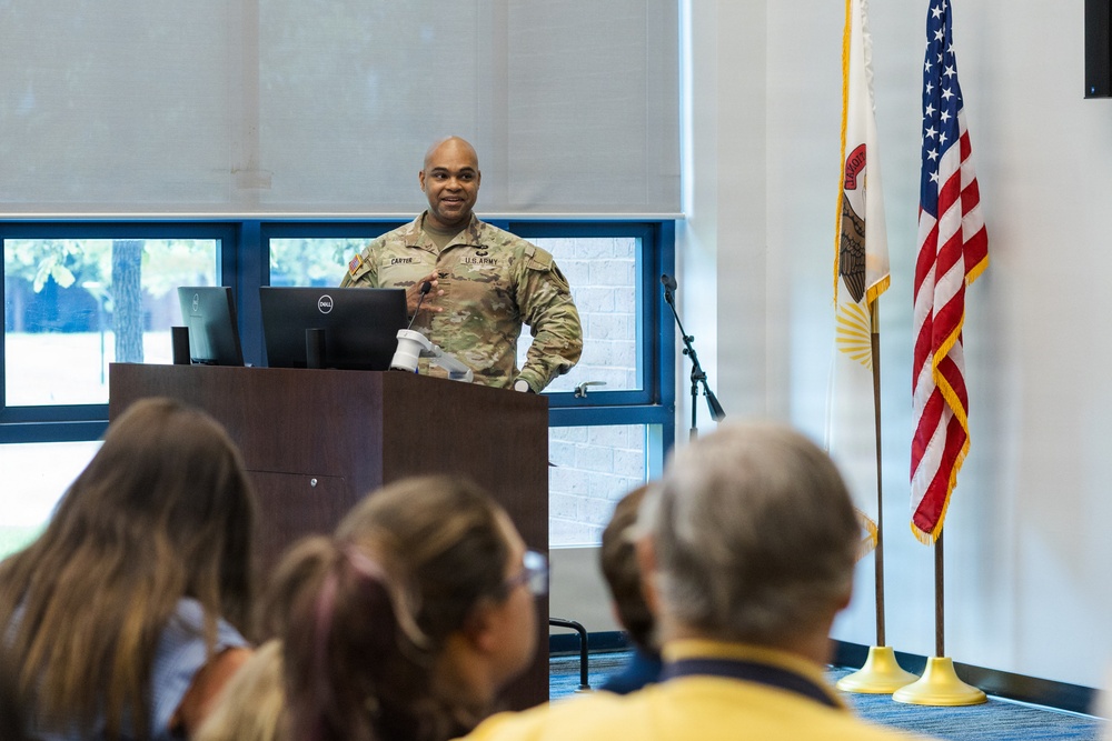 A U.S. Army soldier, identified by name tape as 'CARTER,' is delivering a presentation or briefing at a wooden podium equipped with a Dell monitor. The setting appears to be a formal meeting room or conference hall, with an audience seated in the foreground. Two flags — the United States flag and what appears to be a state flag — are displayed to the right of the speaker.