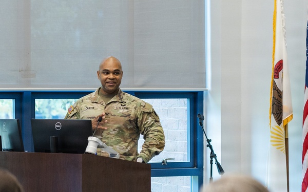 A U.S. Army soldier, identified by name tape as 'CARTER,' is delivering a presentation or briefing at a wooden podium equipped with a Dell monitor. The setting appears to be a formal meeting room or conference hall, with an audience seated in the foreground. Two flags — the United States flag and what appears to be a state flag — are displayed to the right of the speaker.