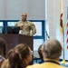 A U.S. Army soldier, identified by name tape as 'CARTER,' is delivering a presentation or briefing at a wooden podium equipped with a Dell monitor. The setting appears to be a formal meeting room or conference hall, with an audience seated in the foreground. Two flags — the United States flag and what appears to be a state flag — are displayed to the right of the speaker.