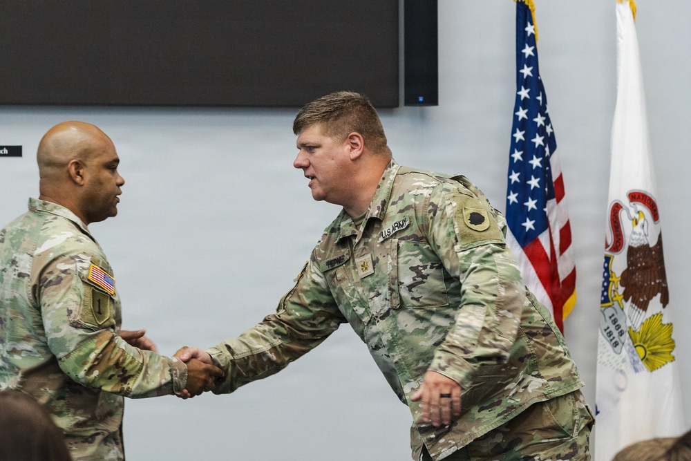 Two U.S. Army soldiers in Operational Camouflage Pattern (OCP) uniforms are shaking hands during what appears to be a formal military ceremony or recognition event. The soldier on the right displays a Major rank insignia (gold oak leaf) on his chest. The setting includes an American flag and what appears to be an Illinois National Guard flag in the background.