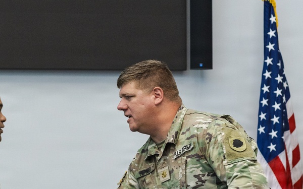 Two U.S. Army soldiers in Operational Camouflage Pattern (OCP) uniforms are shaking hands during what appears to be a formal military ceremony or recognition event. The soldier on the right displays a Major rank insignia (gold oak leaf) on his chest. The setting includes an American flag and what appears to be an Illinois National Guard flag in the background.