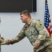 Two U.S. Army soldiers in Operational Camouflage Pattern (OCP) uniforms are shaking hands during what appears to be a formal military ceremony or recognition event. The soldier on the right displays a Major rank insignia (gold oak leaf) on his chest. The setting includes an American flag and what appears to be an Illinois National Guard flag in the background.