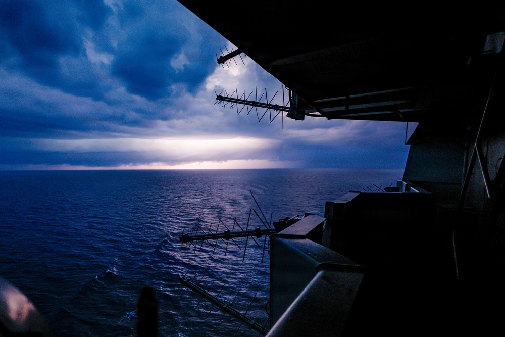 A view from the deck or superstructure of a large naval or military vessel at dusk or dawn, looking out over a calm open ocean. Multiple antenna arrays are visible in silhouette against a dramatic sky with storm clouds and a bright horizon break. The image is taken from an elevated vantage point, likely a bridge wing or observation deck.