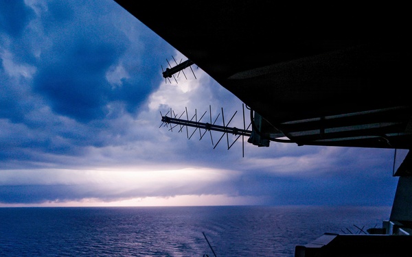 A view from the deck or superstructure of a large naval or military vessel at dusk or dawn, looking out over a calm open ocean. Multiple antenna arrays are visible in silhouette against a dramatic sky with storm clouds and a bright horizon break. The image is taken from an elevated vantage point, likely a bridge wing or observation deck.