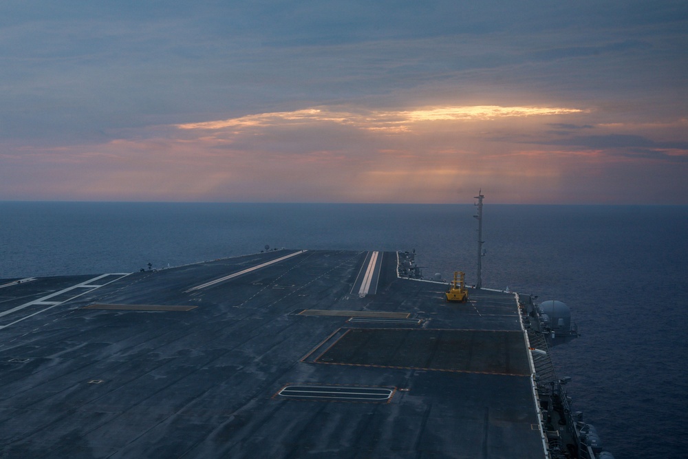 An aerial view of a large aircraft carrier flight deck photographed at dusk or dawn, with the deck empty of aircraft. The carrier is underway at sea, with calm open ocean visible on both sides and a dramatic sky with orange-tinted clouds on the horizon. The perspective appears to be taken from the ship's island superstructure looking forward along the flight deck.