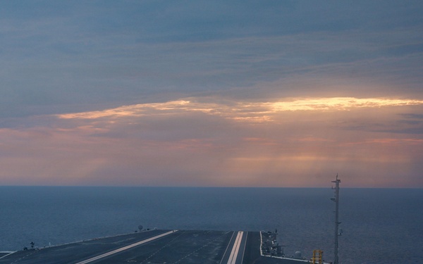 An aerial view of a large aircraft carrier flight deck photographed at dusk or dawn, with the deck empty of aircraft. The carrier is underway at sea, with calm open ocean visible on both sides and a dramatic sky with orange-tinted clouds on the horizon. The perspective appears to be taken from the ship's island superstructure looking forward along the flight deck.