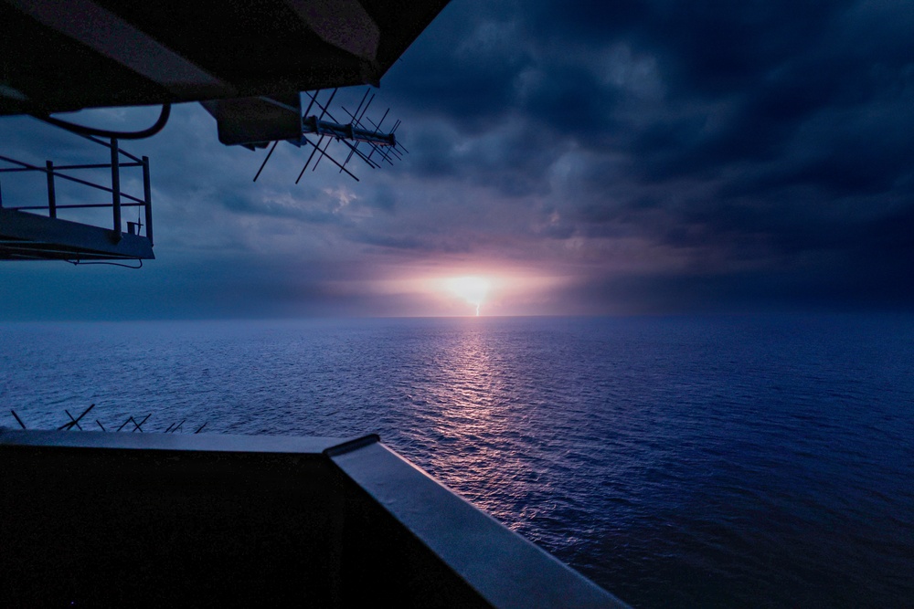 A dramatic seascape photographed from the deck or bridge wing of a large vessel, showing a glowing light source on the horizon — likely a sunset or distant lightning strike — reflecting across calm open ocean waters. The scene is dominated by deep blue tones with heavy storm clouds overhead and a concentrated bright light at the horizon center. Ship infrastructure including antennas, railings, and structural platforms are visible in the upper-left foreground.