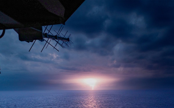 A dramatic seascape photographed from the deck or bridge wing of a large vessel, showing a glowing light source on the horizon — likely a sunset or distant lightning strike — reflecting across calm open ocean waters. The scene is dominated by deep blue tones with heavy storm clouds overhead and a concentrated bright light at the horizon center. Ship infrastructure including antennas, railings, and structural platforms are visible in the upper-left foreground.
