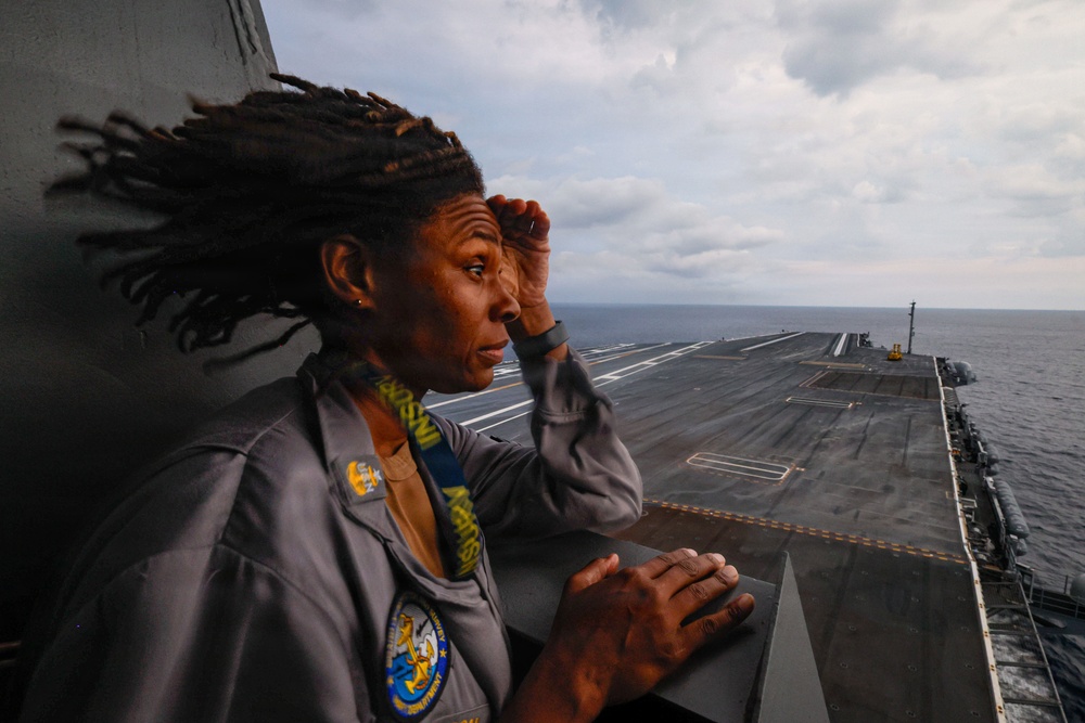 A Navy service member stands on an elevated observation position aboard what appears to be a large aircraft carrier at sea, shielding their eyes while looking out over the flight deck. The individual is wearing a gray Navy uniform with visible patches and insignia, with dreadlocks blowing in the wind. The flight deck stretches into the background with the open ocean visible on the horizon under a partly cloudy sky.