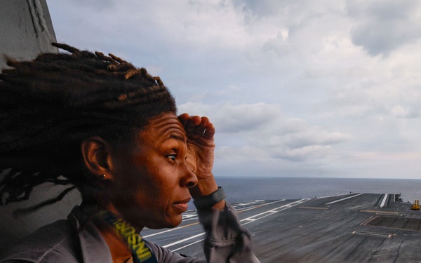 A Navy service member stands on an elevated observation position aboard what appears to be a large aircraft carrier at sea, shielding their eyes while looking out over the flight deck. The individual is wearing a gray Navy uniform with visible patches and insignia, with dreadlocks blowing in the wind. The flight deck stretches into the background with the open ocean visible on the horizon under a partly cloudy sky.