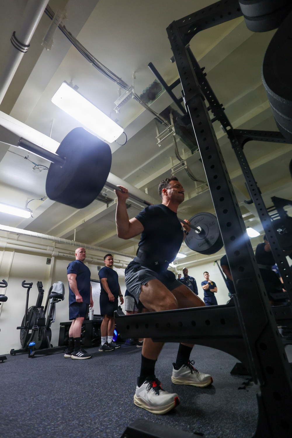 A person in a navy blue t-shirt and shorts is performing a barbell overhead press or push press inside an indoor gym facility. Several observers dressed in matching dark blue uniforms stand in the background watching. The gym appears to be a compact, institutional fitness space with exposed ceiling pipes and fluorescent lighting.
