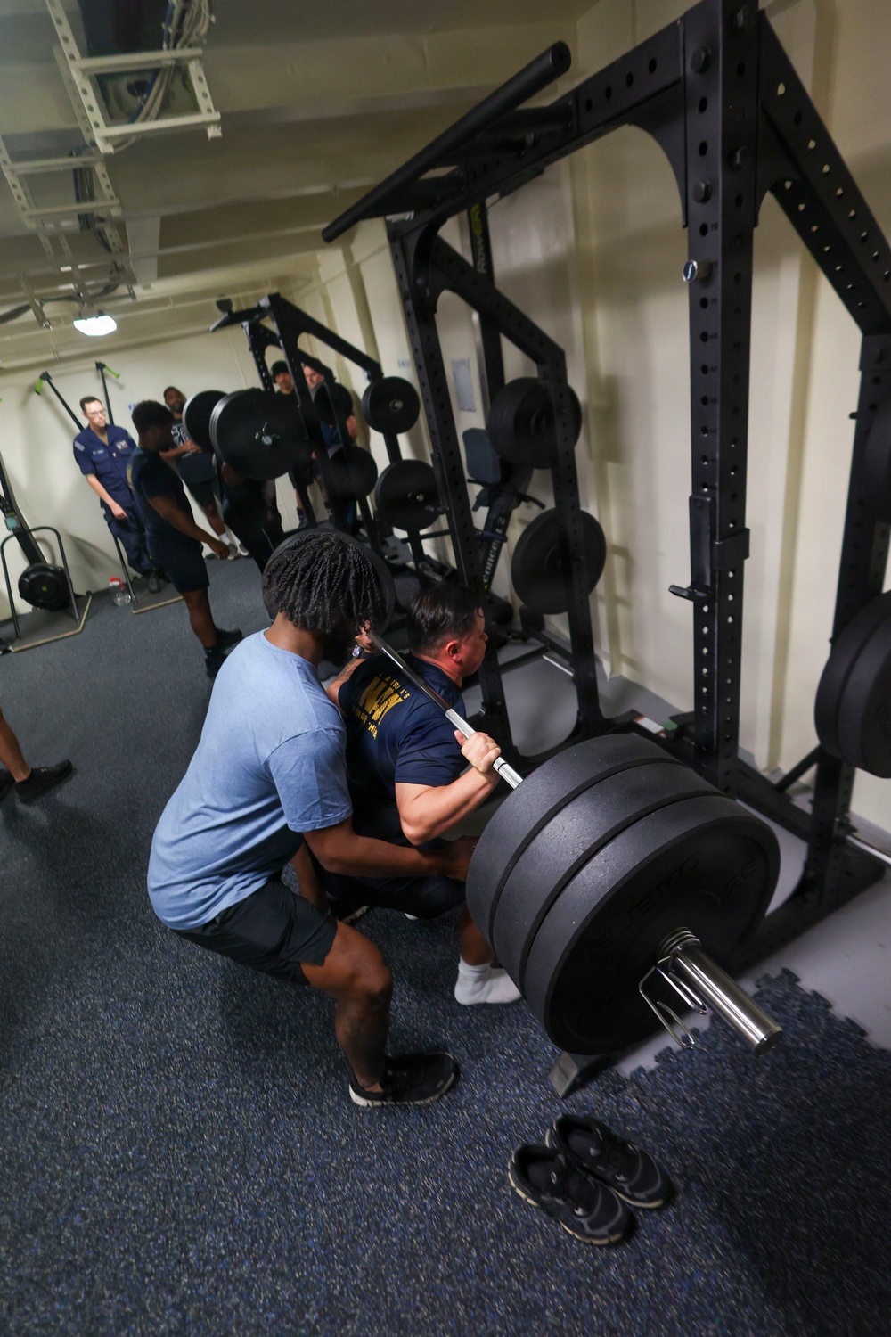 A gym setting where one person is performing a barbell back squat with heavy weight plates loaded on the bar, while a spotter assists from behind. Several other individuals are visible in the background, some observing and others near additional power racks. The facility appears to be an indoor weight room with multiple squat/power racks and rubber flooring.