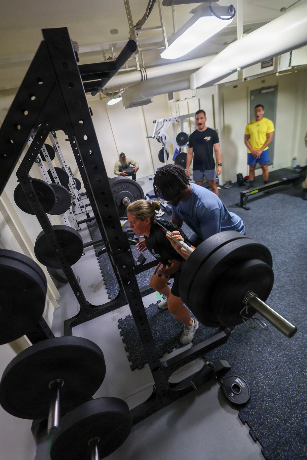 A group of people are working out in a compact weight room, likely aboard a military vessel or institutional facility. A woman in a black shirt is performing a deadlift or bent-over row with a heavily loaded barbell while a spotter in a blue shirt assists her. Several other individuals are visible in the background, with two men in black and yellow shirts observing or reacting with apparent surprise or encouragement.