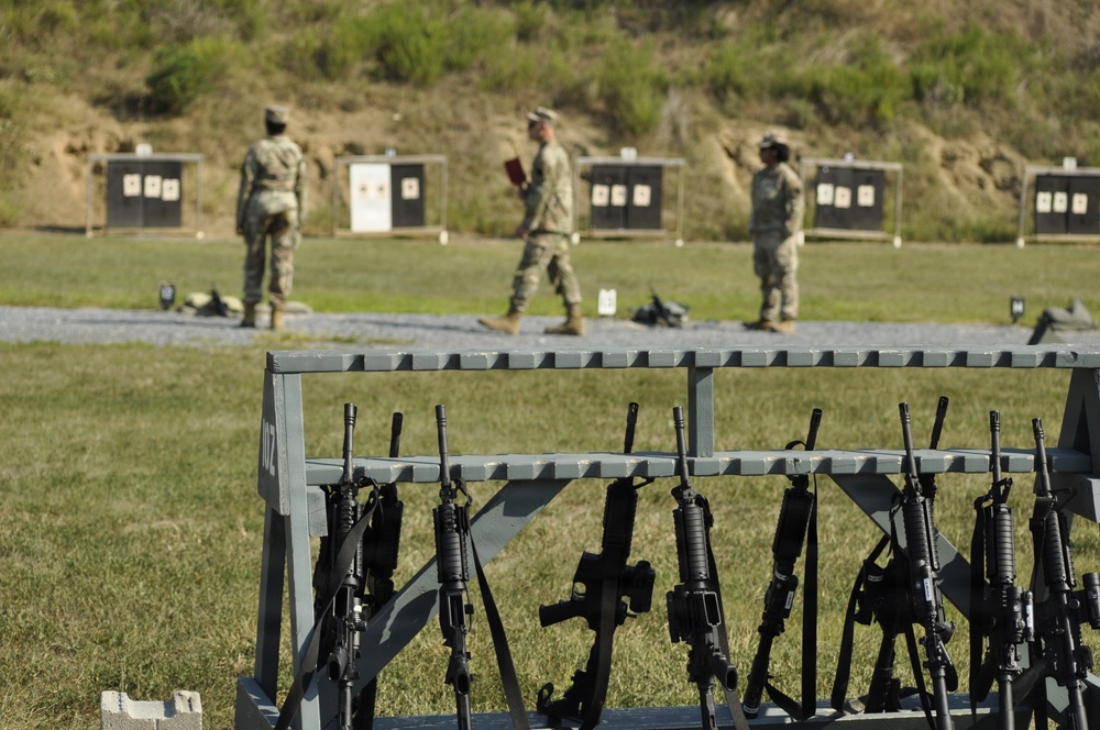 A military firing range with multiple rifles secured in a foreground weapon rack. Three soldiers in camouflage uniforms are visible in the background, appearing to inspect or score targets. The range features multiple black silhouette targets and one white paper target mounted on stands.