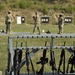 A military firing range with multiple rifles secured in a foreground weapon rack. Three soldiers in camouflage uniforms are visible in the background, appearing to inspect or score targets. The range features multiple black silhouette targets and one white paper target mounted on stands.