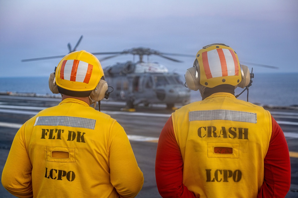 Two naval personnel wearing yellow safety vests and helmets with hearing protection stand on a flight deck observing a military helicopter. The individual on the left is identified as 'FLT DK LCPO' (Flight Deck Leading Chief Petty Officer) and the one on the right as 'CRASH LCPO' (Crash and Salvage Leading Chief Petty Officer). The scene appears to be aboard a naval vessel at dusk or dawn, with open ocean visible in the background.