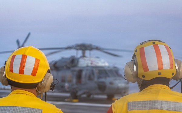 Two naval personnel wearing yellow safety vests and helmets with hearing protection stand on a flight deck observing a military helicopter. The individual on the left is identified as 'FLT DK LCPO' (Flight Deck Leading Chief Petty Officer) and the one on the right as 'CRASH LCPO' (Crash and Salvage Leading Chief Petty Officer). The scene appears to be aboard a naval vessel at dusk or dawn, with open ocean visible in the background.
