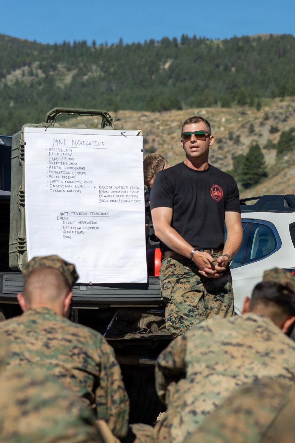 A military instructor in a black t-shirt and camouflage pants conducts an outdoor field briefing on mountain navigation and anti-tracking techniques for a group of Marines in MARPAT camouflage uniforms. The briefing is taking place in a mountainous, forested environment with a whiteboard mounted on a military vehicle displaying handwritten notes. The setting appears to be a field training exercise.