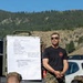 A military instructor in a black t-shirt and camouflage pants conducts an outdoor field briefing on mountain navigation and anti-tracking techniques for a group of Marines in MARPAT camouflage uniforms. The briefing is taking place in a mountainous, forested environment with a whiteboard mounted on a military vehicle displaying handwritten notes. The setting appears to be a field training exercise.
