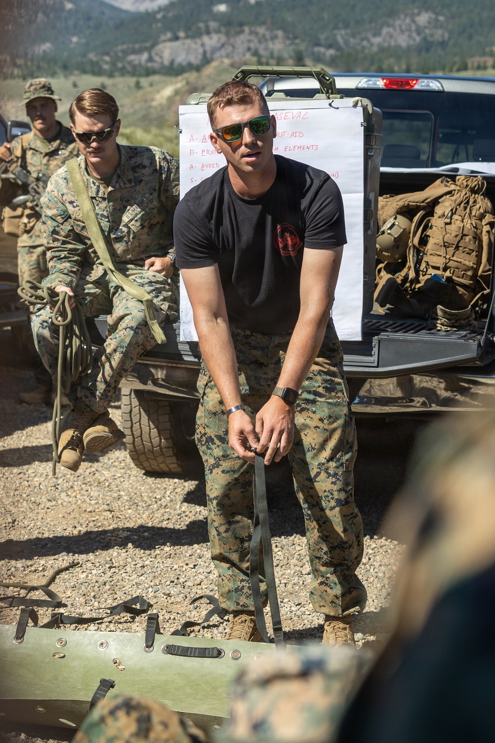 A military training exercise is taking place outdoors in a mountainous terrain. A man in a black t-shirt and MARPAT camouflage pants appears to be an instructor demonstrating casualty evacuation (CASEVAC) procedures using a litter/stretcher. Several uniformed Marines in full digital camouflage are observing and participating in the training.