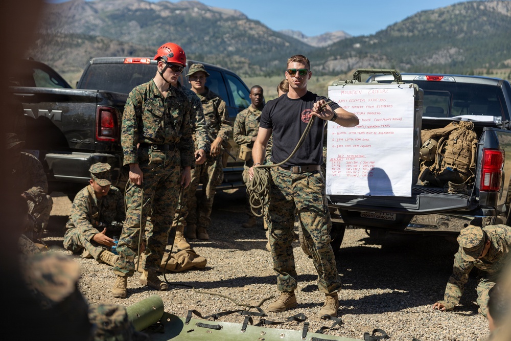 A military training session is taking place outdoors in a mountainous environment. An instructor in a black t-shirt is demonstrating rope techniques to a group of personnel in camouflage uniforms, with a whiteboard displaying 'Mountain Casualty' training notes mounted on a pickup truck. The setting appears to be a field training exercise focused on mountain casualty care and handling procedures.