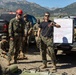 A military training session is taking place outdoors in a mountainous environment. An instructor in a black t-shirt is demonstrating rope techniques to a group of personnel in camouflage uniforms, with a whiteboard displaying 'Mountain Casualty' training notes mounted on a pickup truck. The setting appears to be a field training exercise focused on mountain casualty care and handling procedures.