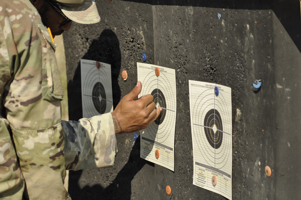 A soldier in MultiCam/OCP camouflage uniform is examining shooting targets posted on a dark backstop board at a firing range. The individual appears to be scoring or marking bullet holes on the paper targets. Three circular bullseye targets are visible, pinned to the board with colored pushpins.