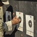 A soldier in MultiCam/OCP camouflage uniform is examining shooting targets posted on a dark backstop board at a firing range. The individual appears to be scoring or marking bullet holes on the paper targets. Three circular bullseye targets are visible, pinned to the board with colored pushpins.
